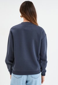 Marineblauwe sweatshirt met een ronde hals, geribbelde mouwen en zoom. Zachte textuur, minimalistisch ontwerp en een vierkante patch op de middelste achterkant.