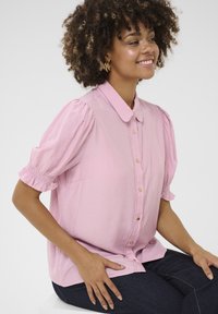 Svetloružová blúzka s krátkym rukávom a klasickým golierom, s predným zapínaním na gombíky a nariadenými pufovými rukávmi. Obsahuje dekoratívne šitie pozdĺž zapínania.