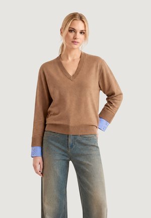 Mujer con cabello rubio que lleva un suéter marrón de cuello en V sobre una camisa azul, combinado con vaqueros azules desgastados, de pie frente a un fondo liso.