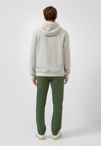 Mężczyzna w teksturowanym jasnoszarym swetrze z kapturem, zielonych spodniach i białych trampkach stoi przodem do gładkiej białej ściany.