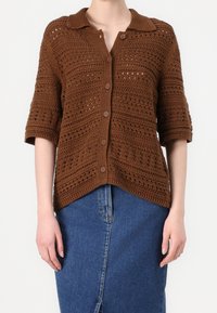 Cardigan marrón de punto con cuello, mangas cortas y botones al frente. Presenta patrones de enrejado abierto y detalles acanalados en las mangas y el dobladillo.