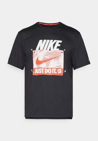 Černé bavlněné tričko s velkým bílým logem "NIKE" a grafickým motivem "JUST DO IT" v červené a bílé barvě, doplněné texturou a kovovými oky.