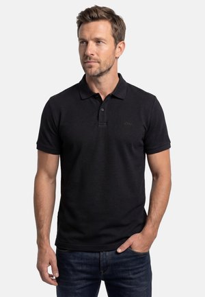 Uomo con una polo nera a maniche corte e jeans blu, in piedi con una mano in tasca, che guarda leggermente di lato.