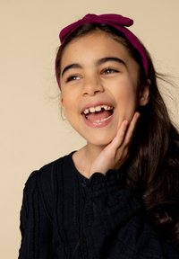 Kind dat een marineblauwe gestructureerde trui met gerimpelde manchetten en een fuchsia hoofdband draagt, terwijl ze glimlacht met een hand op haar wang tegen een beige achtergrond.