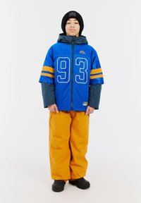 Blaue sportliche Jacke mit der Nummer 93, orangefarbene Streifen, kombiniert mit orangefarbenen wasserdichten Hosen und schwarzen Schuhen. Tragen mit einer schwarzen Mütze.