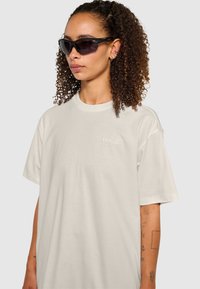 Lys beige oversized t-shirt med korte ærmer, prydet med et lille, broderet "HALO" logo på brystet. Model iført mørke solbriller.