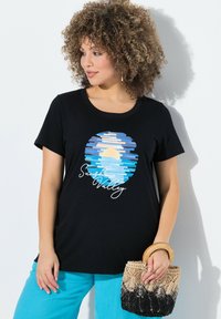 Černé bavlněné tričko s kulatým potiskem s modrým a žlutým designem západu slunce, spárované se světle modrými kalhotami a tkanou kabelkou.