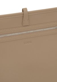 Bolso de cuero beige con una textura suave, con cierre superior de cremallera y logo "TOUS" en relieve. Presenta un detalle destacado de cremallera.