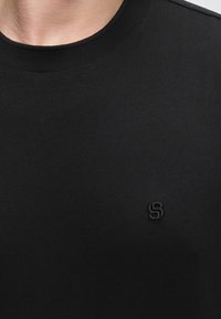 T-shirt noir à col rond porté par une personne avec un petit logo embossé « SB » sur le côté gauche de la poitrine.