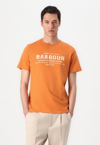 T-shirt de algodão laranja com decote redondo, apresentando texto branco: "BARBOUR QUALITY OUTDOOR APPAREL" e detalhes de fundação abaixo.