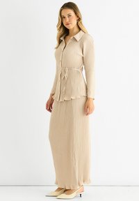 Beige ribbed dress with a collar, long sleeves, and a waist tie. Features a flared bottom edge and is paired with light-colored heeled shoes.