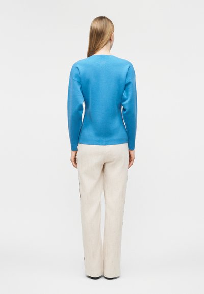 Γυναίκα που στέκεται γυρισμένη προς τα πίσω, φορώντας ένα φωτεινό μπλε μακρυμάνικο τοπ και φαρδιά μπεζ παντελόνια μπροστά σε απλό φόντο.