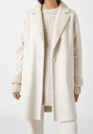 Femme portant un long manteau crème avec une doublure sherpa au col et aux poignets, sur un pull en maille texturée et un pantalon en maille côtelée.