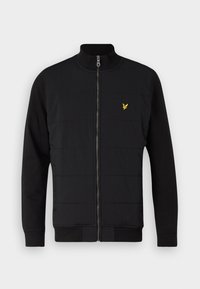 Giacca nera con zip frontale, caratterizzata da un design trapuntato sul corpo, maniche lunghe lisce e un piccolo logo giallo sul petto.