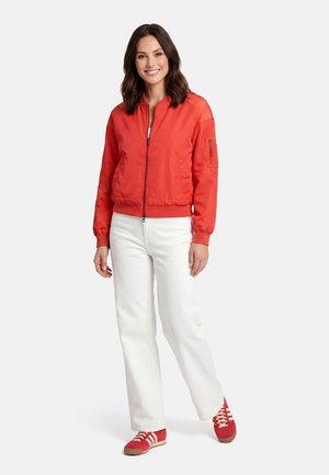 Vrouw die lacht, draagt een rode jas met ritssluiting, witte broek en rode sneakers, staand tegen een effen lichte achtergrond.