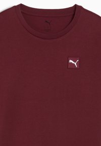 Bordeaux crew neck t-shirt lavet af blødt bomuldsstof, med et hvidt Puma logo patch på venstre brystområde. Glat tekstur uden mønstre.