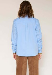 Chemise bleu clair à manches longues avec un pli au dos et des poignets. Associée à un pantalon en velours côtelé marron avec un effet de côtes verticales.
