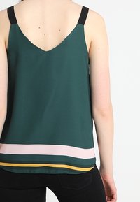 Zelený bezramenný top s černými ramínky, ve tvaru V na zádech a výraznými horizontálními pruhy v růžové, tmavě zelené a žluté barvě na spodním lemu.