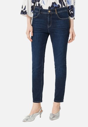 Femme portant un jean slim bleu foncé avec cinq poches, fermeture à boutons et zip, associé à des chaussures à talons bas argentées avec soutien au talon.