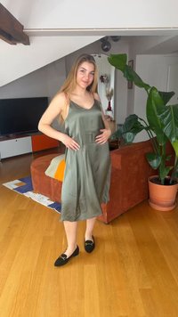 Νεαρή γυναίκα με ελιά πράσινο φόρεμα και μαύρα μπαλαρίνια στέκεται σε ένα σαλόνι κοντά σε ένα μεγάλο φυτωριό και σε ένα καφέ σκουριάς καναπέ.