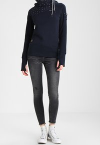 Sudadera con capucha azul marino de textura en la parte superior, contrastada con una capucha estampada, combinada con jeans ajustados grises y zapatillas blancas.