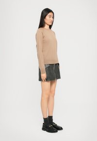 Kvinne som står sidelangs iført en beige genser, svart skinn mini skjørt med glidelås foran, mørke sokker og svarte loafers mot hvit bakgrunn.
