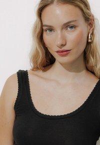 Débardeur noir en maille côtelée avec un col festonné. Le modèle porte des boucles d'oreilles en forme de goutte d'or. Peau lisse avec un maquillage naturel ; cheveux ondulés.