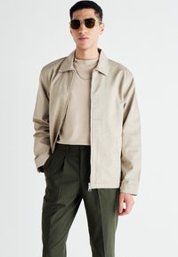 Veste beige à fermeture éclair avec un col plat, portée sur une chemise beige clair et un pantalon vert foncé. Présente un design épuré et un matériel minimal.