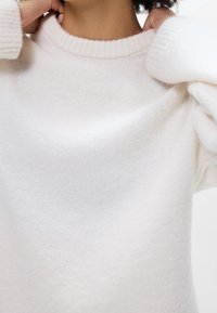 Personne ajustant le col côtelé d'un pull blanc doux à manches longues avec une texture duveteuse.
