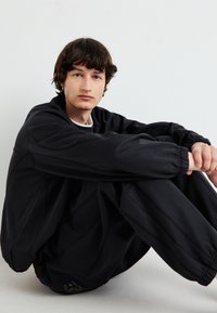 Hanorac supradimensionat negru și pantaloni de trening asortați, cu manșete elastice și un finisaj texturat. Modelul este așezat pe un fundal simplu.