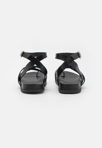 Schwarze flache Sandalen verfügen über eine strukturierte Gummisohle und überkreuzte synthetische Riemen mit einem verstellbaren Schnallenverschluss am Knöchel.