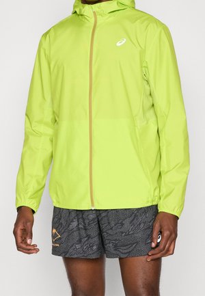 Homme portant une veste à capuche zippée vert néon et un short de sport noir à motifs, debout devant un fond uni.