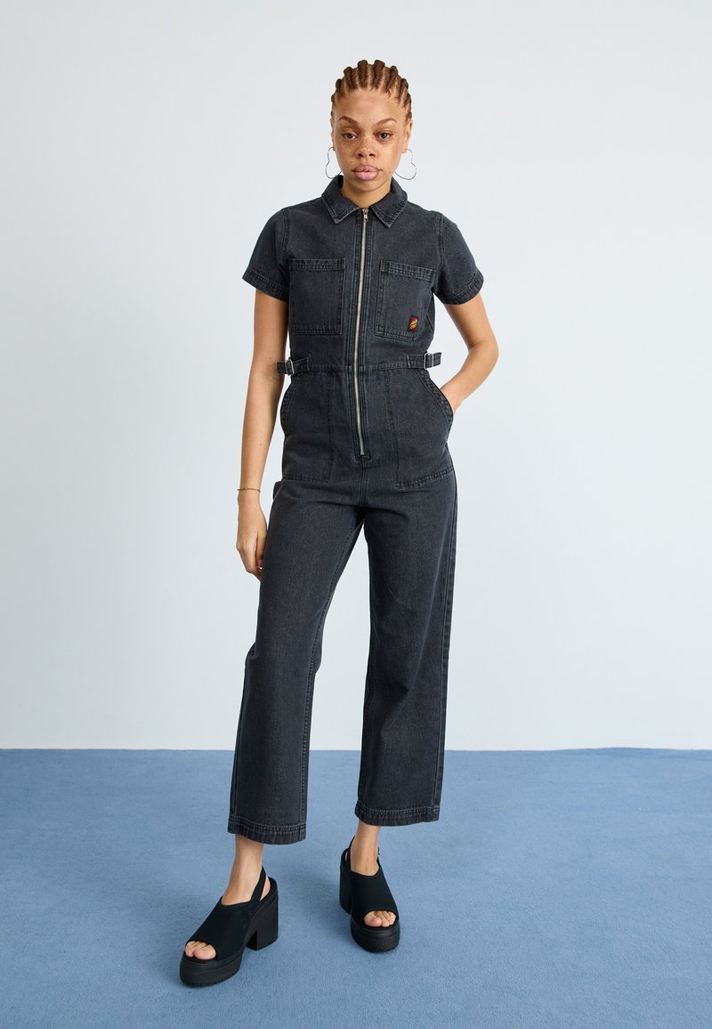 Sorte denim jumpsuit med kortermet, glidelås foran, to lommer og belte i midjen. Stylet med svarte platå sko. Ensfarget bakgrunn.