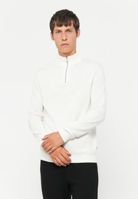 Weißer Pullover mit Reißverschluss am Hals, langen Ärmeln, gerippten Bündchen und Saum; weiche Textur und minimalistisches Design. Schwarze Hose nicht enthalten.