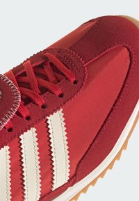 Červená športová obuv s textúrovaným zvrškom zo semišu, tromi bielymi prúžkami v tvare cikcaku a gumovou podrážkou so vzorom pre lepšiu trakciu.