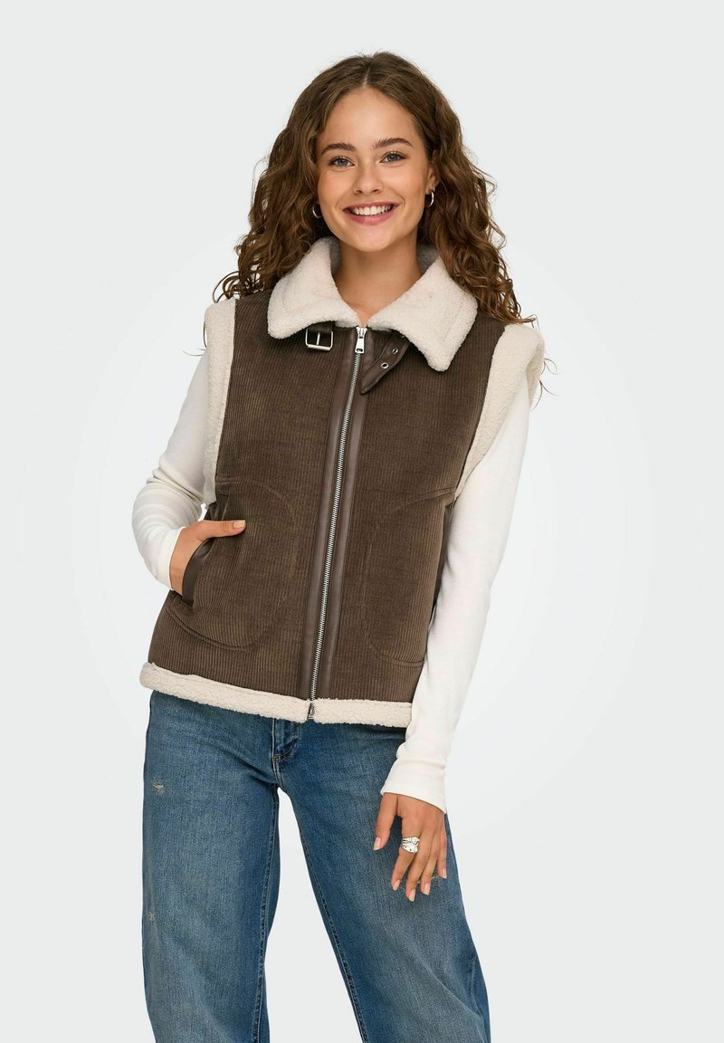 Brun kordfløjl vest med creme sherpa-foring, sølv lynlås og justerbar kravebuckle. Båret over en langærmet skjorte og jeans.