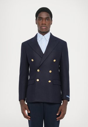Blazer doppiopetto blu navy in lana, con bottoni dorati, revers a scollatura e due tasche a patta. Indossato sopra una camicia a righe blu.