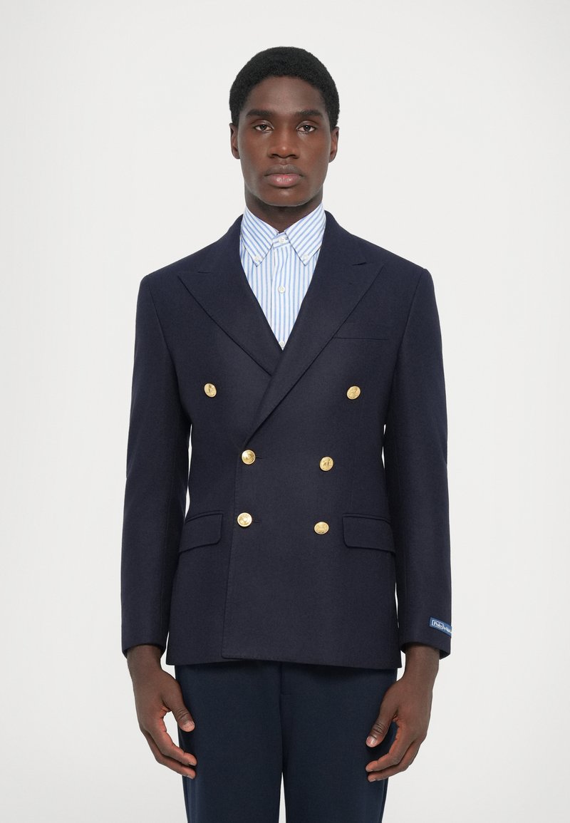 Blazer croisé bleu marine en laine, avec des boutons dorés, des revers crantés et des poches à rabat doubles. Porté par-dessus une chemise rayée bleue.