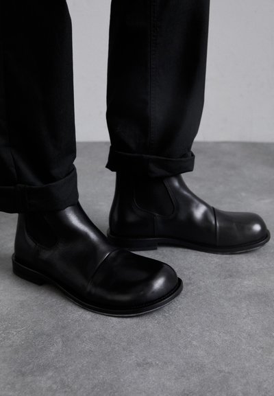 Cizme negre din piele până la glezna, cu vârfuri rotunde, panouri laterale elastice și tălpi plate din cauciuc, purtate cu pantaloni negri rulați pe o suprafață gri.