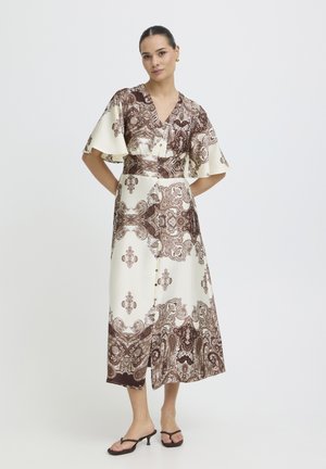 Kobieta stojąca z rękami za plecami, ubrana w kremowo-brązową sukienkę midi z dekoltem w serek i wzorem oraz czarne sandały na obcasie na białym tle.