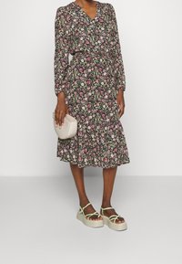 Kukkakuvioinen mekko, jossa pitkät hihat, V-aukko, musta pohja, vaaleanpunaisia ja valkoisia kukkia, röyhelöinen helma. Yhdistetty beigeen laukkuun ja vihreisiin remmikenkiin.