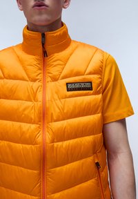 Pomarańczowa kamizelka puchowa z zamkiem błyskawicznym, poziomym pikowaniem, wysokim kołnierzem i wyszywaną naszywką z logo; noszona na pasującym pomarańczowym t-shircie.