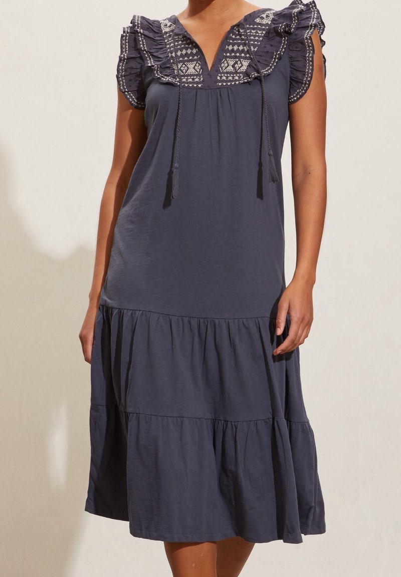 Femme portant une robe bleu foncé sans manches avec des épaules brodées à volants et une jupe à étages, debout devant un fond clair uni.