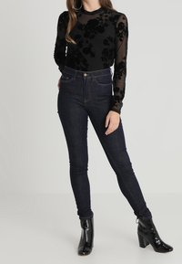 Svart genomskinlig topp med blommönster, i kombination med mörkblå skinny jeans. Modellen bär glansiga svarta ankelstövlar.