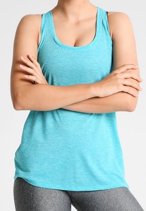 Personne portant un haut sans manches bleu clair et des leggings gris, debout les bras croisés devant un fond uni.