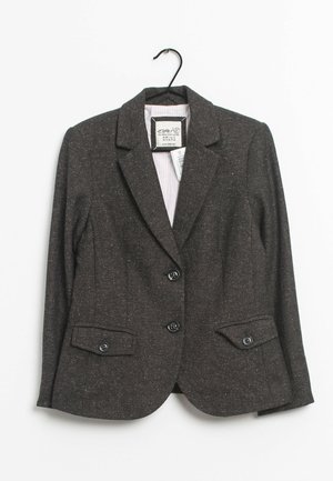 Blazer gris foncé texturé avec deux boutons, deux poches à rabat devant et un col cranté accroché à un cintre noir.