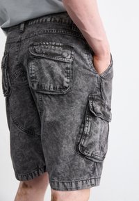 Cargo shorts dalam denim hitam pudar, dilengkapi dengan banyak saku, detail jahitan, dan potongan santai, dikenakan dengan tangan di saku.