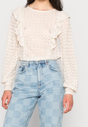 Woman wearing a white lace long-sleeve blouse with ruffles paired with high-waisted light blue checkered jeans.