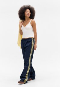 White lace-trimmed camisole, navy joggers with yellow stripes, and a light yellow sweater draped over the shoulder. Brown shoes.