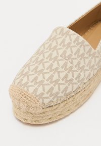 Beige espadrilles ar austa jūtas zoli, ar rakstainu auduma augšdaļu, kas attēlo atkārtotus "MK" logotipus. Klasisks slip-on dizains.